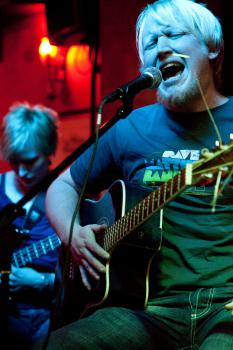 Bandfoto ge$h: Nahaufnahme eines Sängers mit sehr hellblondem Haar, der mit geschlossenen Augen intensiv in ein Mikrofon singt. Er spielt gleichzeitig eine Akustikgitarre. Sein Gesichtsausdruck wirkt emotional und kraftvoll. Im Hintergrund ist unscharf ei